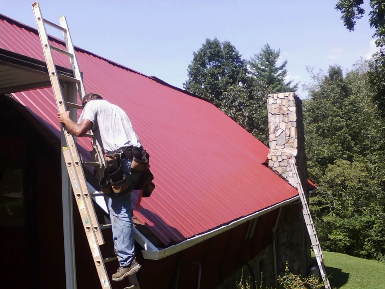 Roofer on a ladder beside a red metal roof and stone chimney — Soffit & Fascia Repair by Roofing Brothers in Catonsville